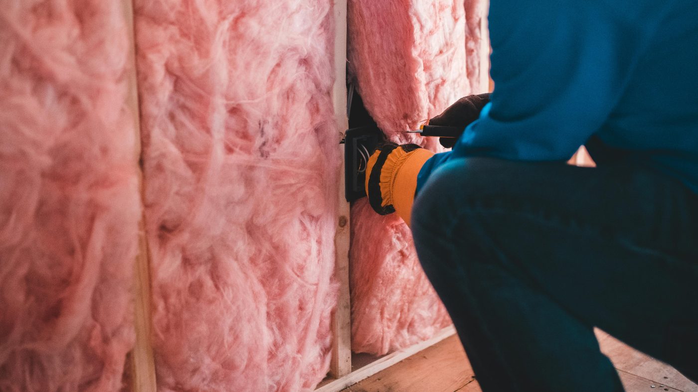 Real construction photo: worker installing pink fiberglass insulation batts in a stud-framed wall cavity — the absorption layer in a sound-proof wall assembly